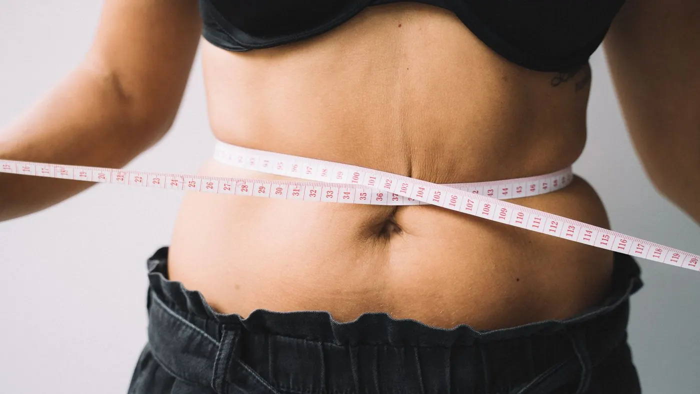 Primer plano del abdomen de una mujer con una cinta métrica rosada rodeando la cintura, sin rostro en el cuadro: la medida de la circunferencia abdominal