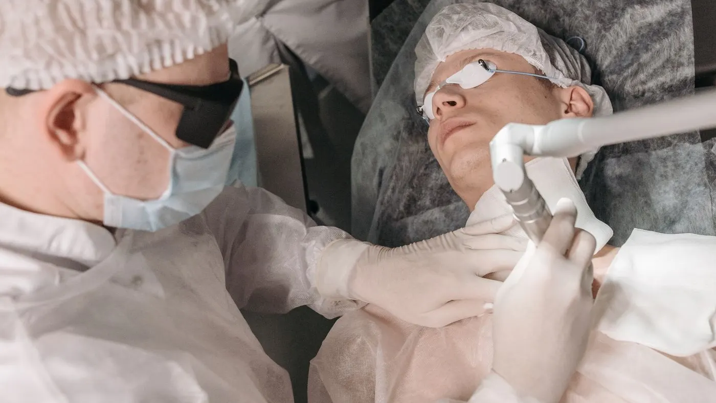 Médico con mascarilla y guantes que aplica un láser sobre la zona del cuello de un paciente con gafas de protección en un consultorio, como en los procedimientos con láser