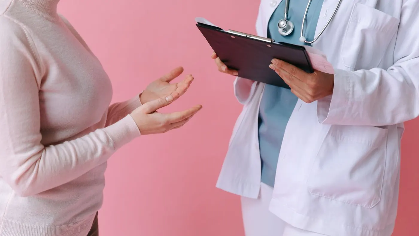 Médica con bata blanca y estetoscopio que sostiene una tablilla mientras una paciente le explica algo con las manos, una consulta de valoración antes de una cirugía como la lipotransferencia