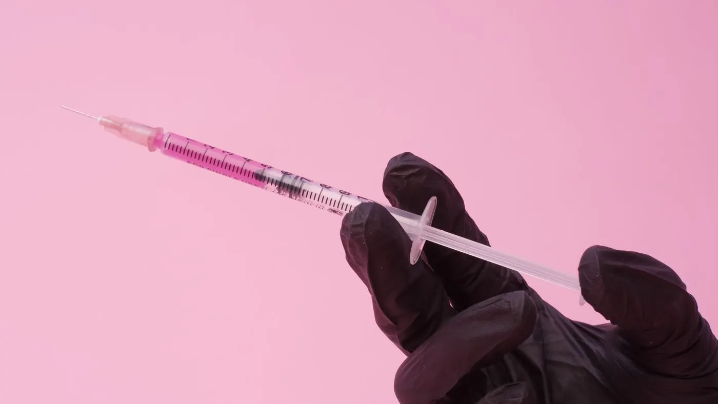 Mano con guante negro que sostiene una jeringa con líquido rosado sobre fondo rosa, imagen de las microinyecciones de la mesoterapia